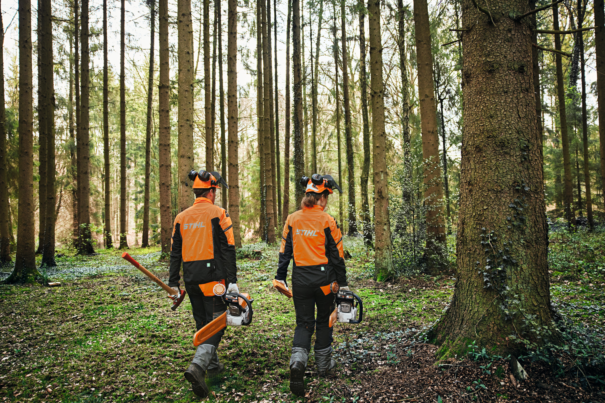 Två personer som bär skyddsutrustning i en skog, med motorsågar och yxa.